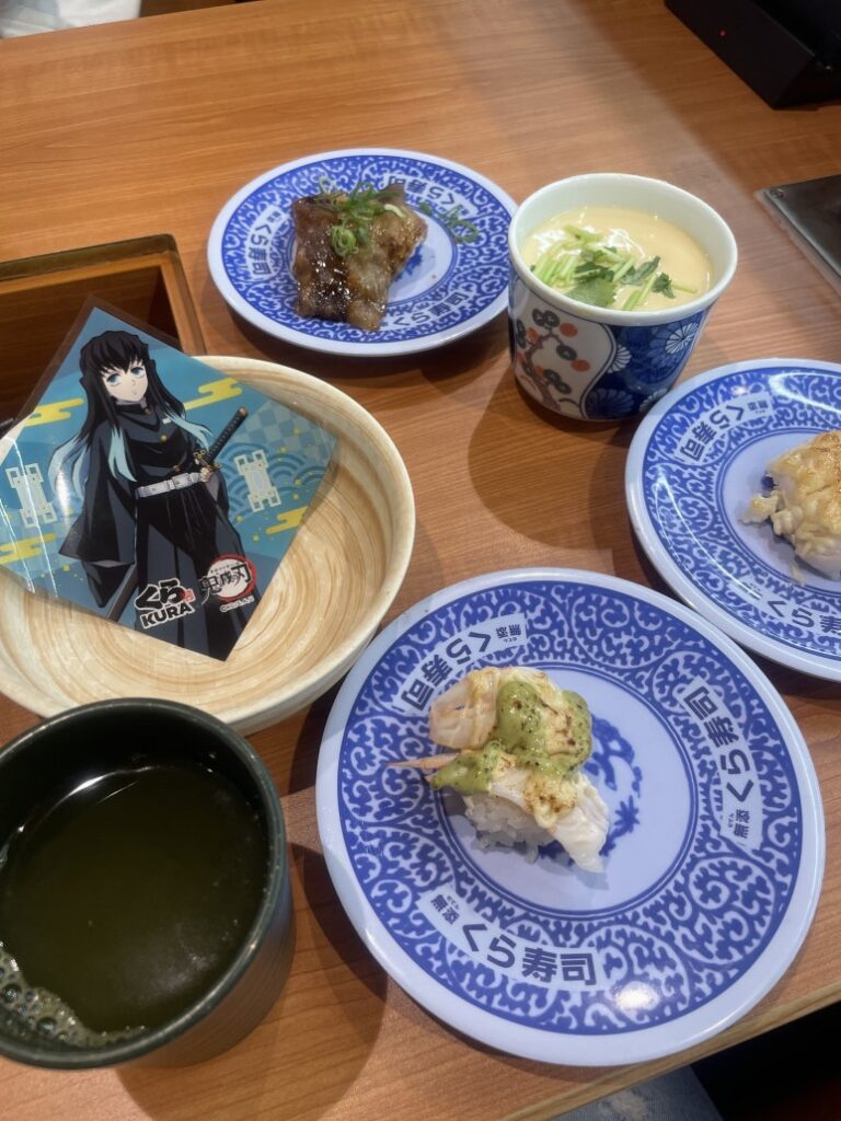 A table with various dishes at Kura Sushi, including sushi, chawanmushi, green tea, and a collectible card
