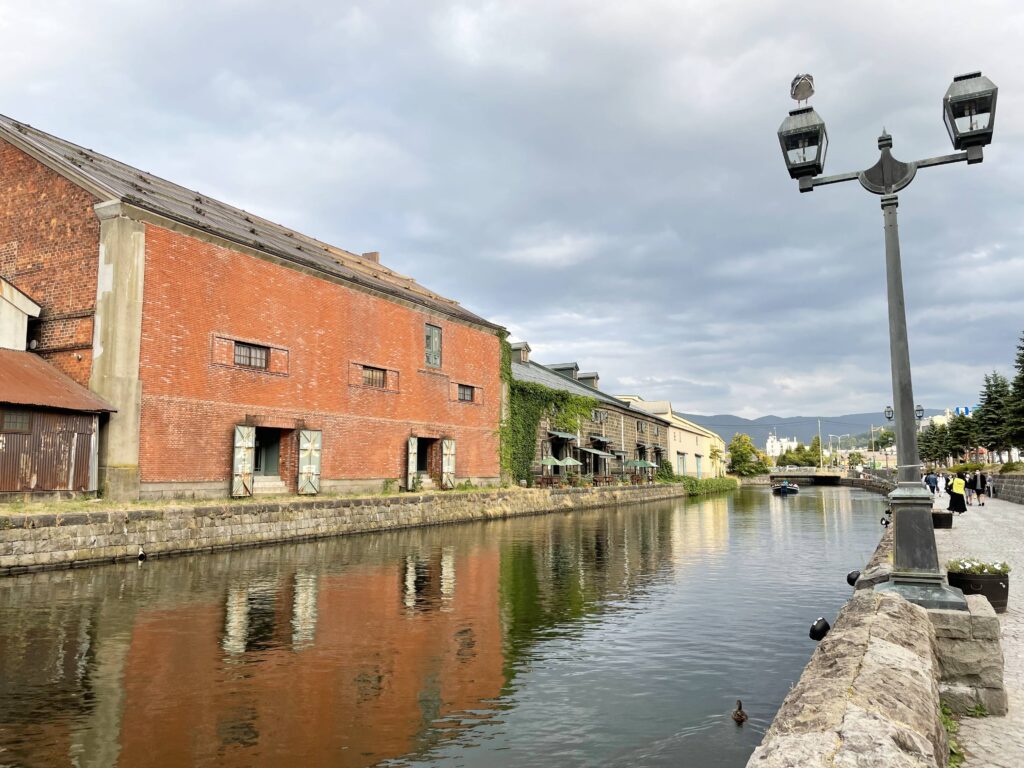 Daytime view of Otaru Canal in Hokkaido, Japan, with historic buildings lining the left side and people strolling or taking photos on the right walkway.