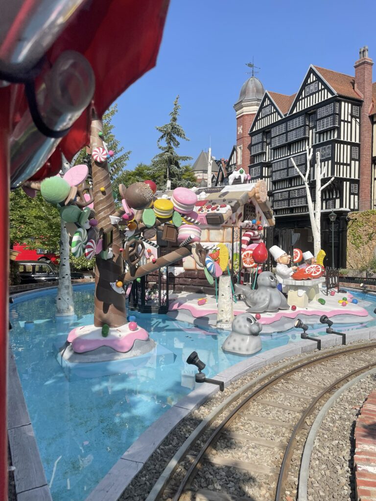 A view from the mini train at Shiroi Koibito Park, showing a candy-themed house and a red brick building in the background.